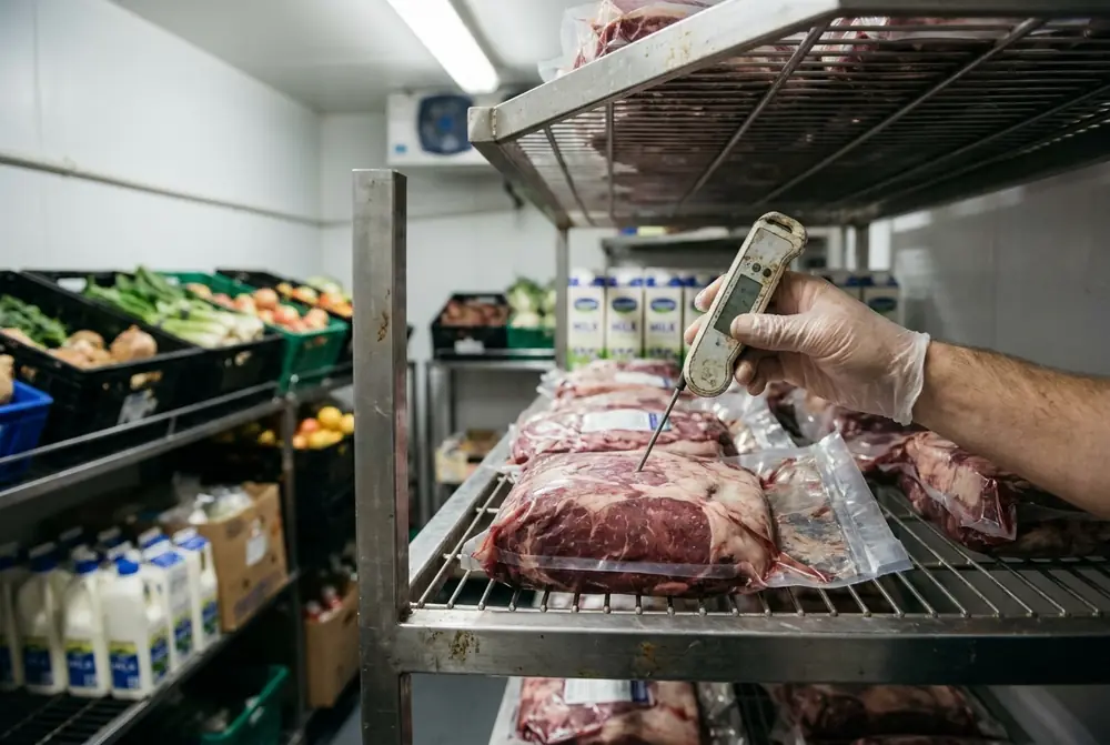 Gloved hand pushing a probe thermometer into vacuum-packed meat on a cool room shelf