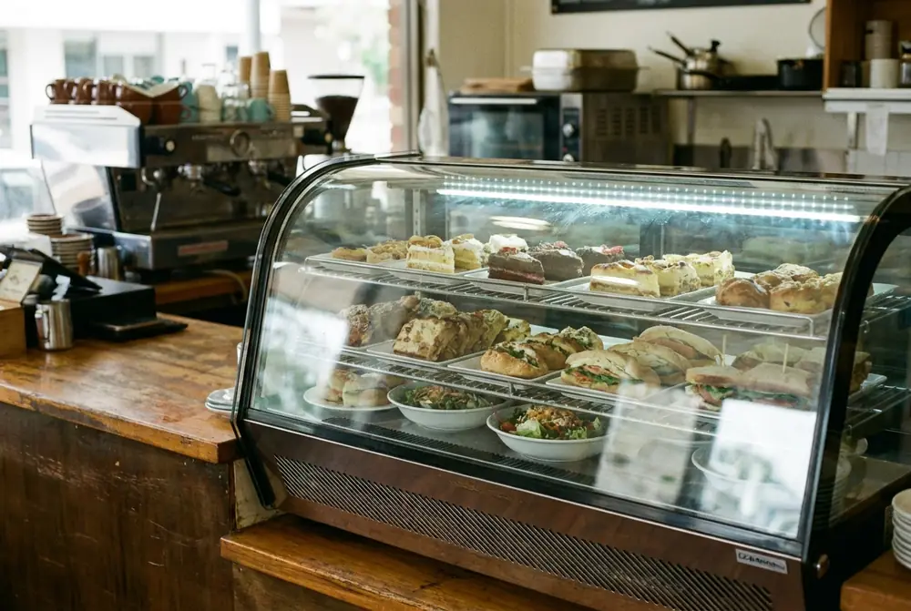 Curved glass cafe display cabinet holding cakes, sandwiches and salads on a timber counter